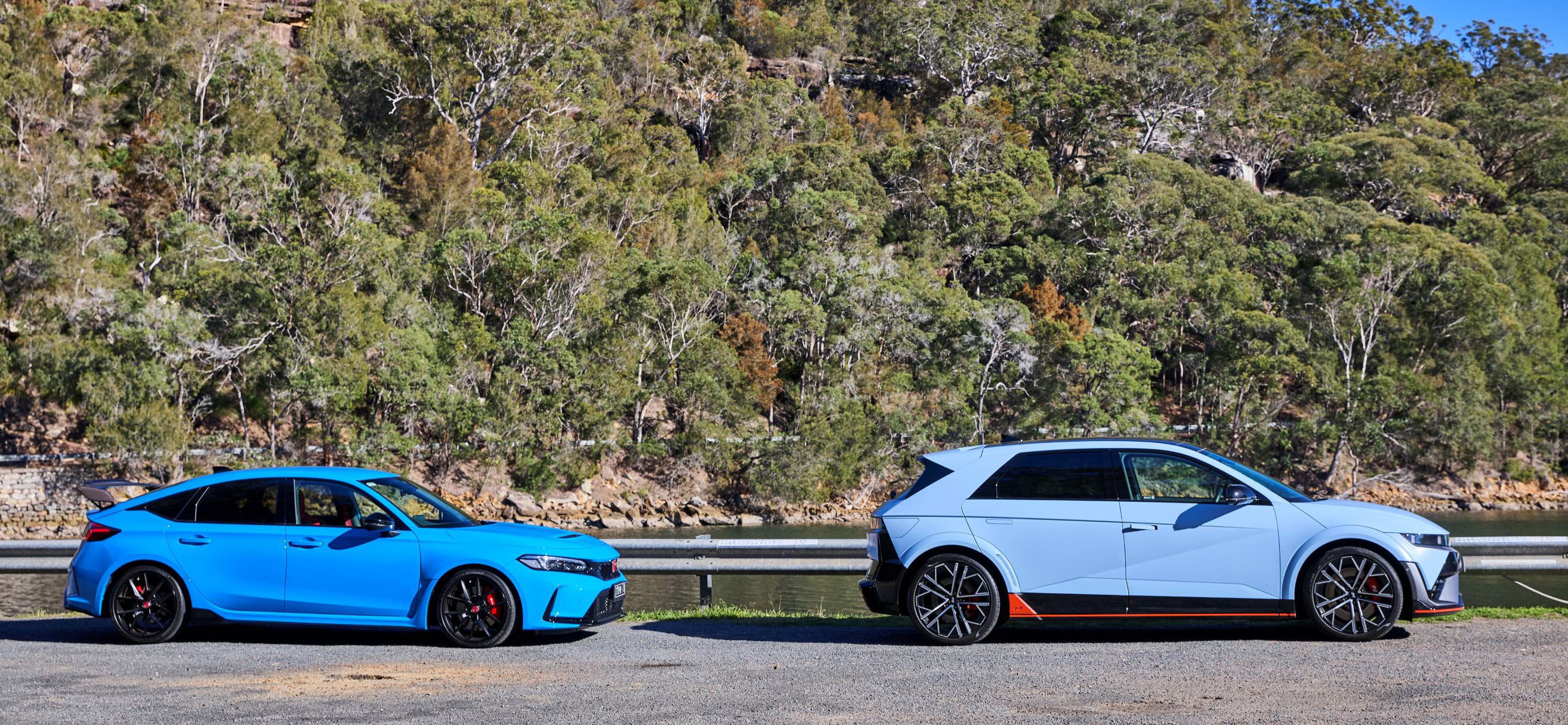 Two blue cars parked side by side along a lakeside road with a dense forested hillside in the background.