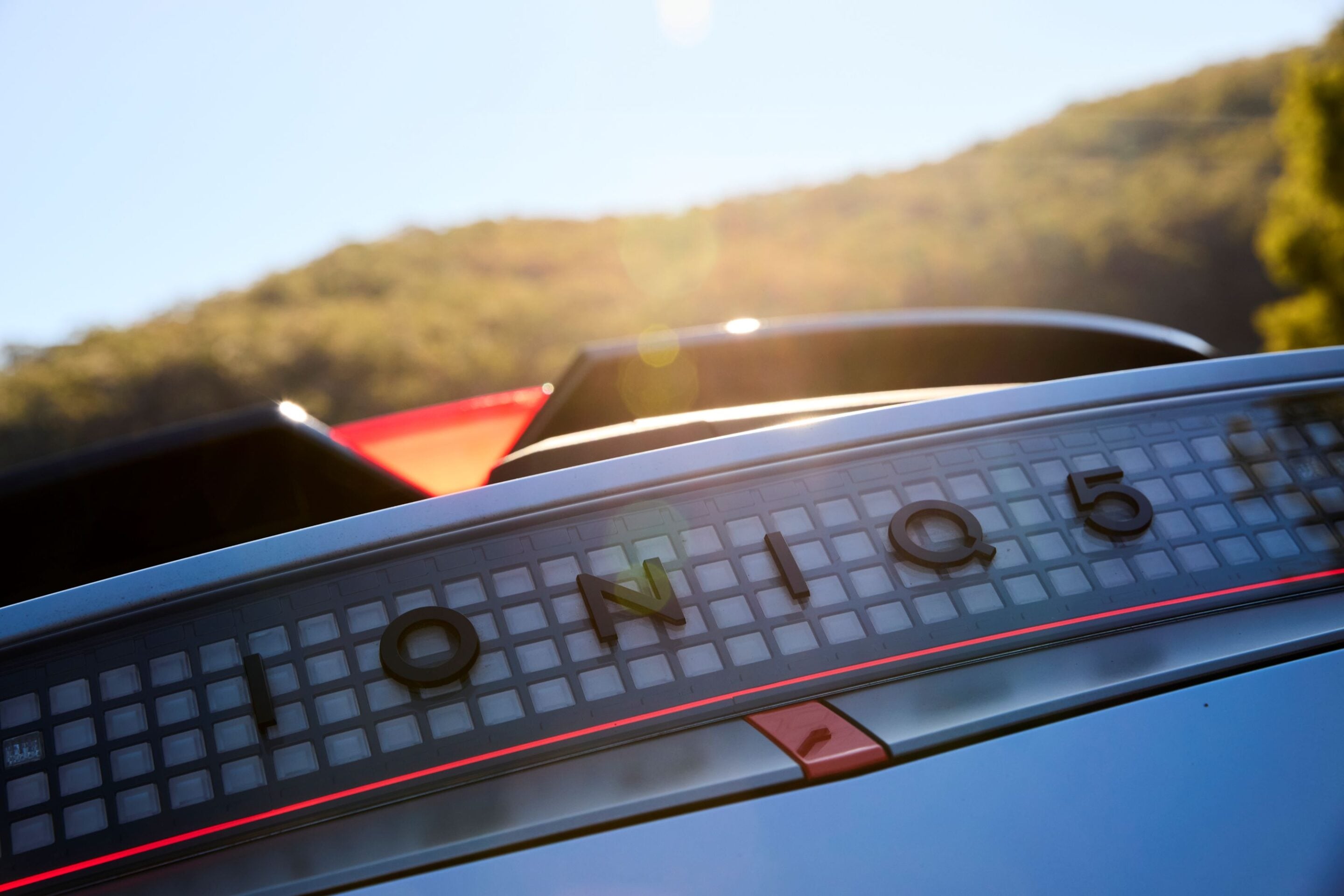 Close-up of a vintage car dashboard gauge with a grid pattern, red accent line, and a blurred hillside and blue sky in the background