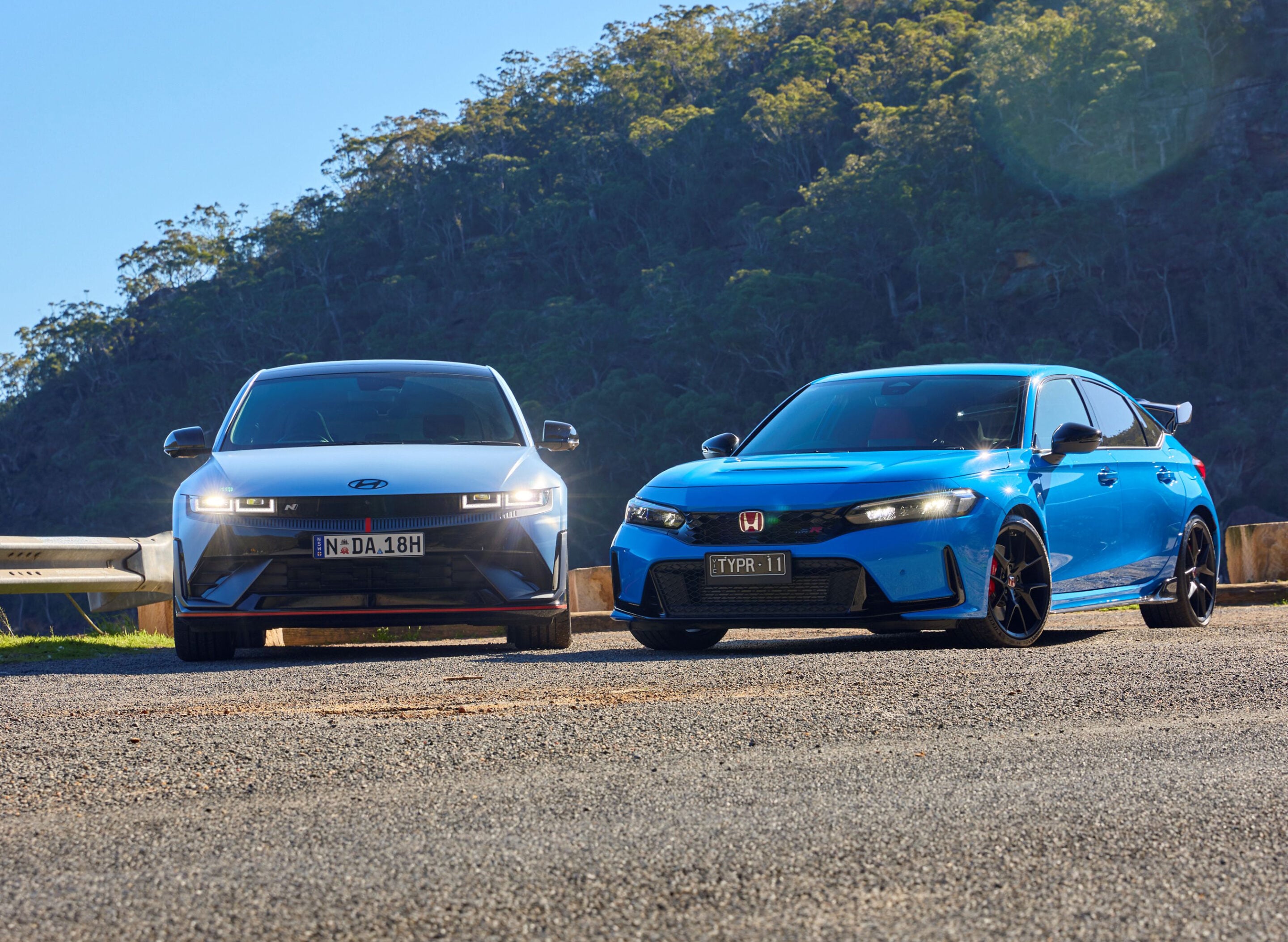 Two cars parked side by side on a sunny day, a white Hyundai on the left and a blue Honda on the right, with a forested hill behind them.