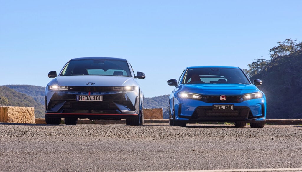 Two modern cars parked side by side on a sunlit road with a clear blue sky behind them. left car is white, right car is blue, with hills in the distance.