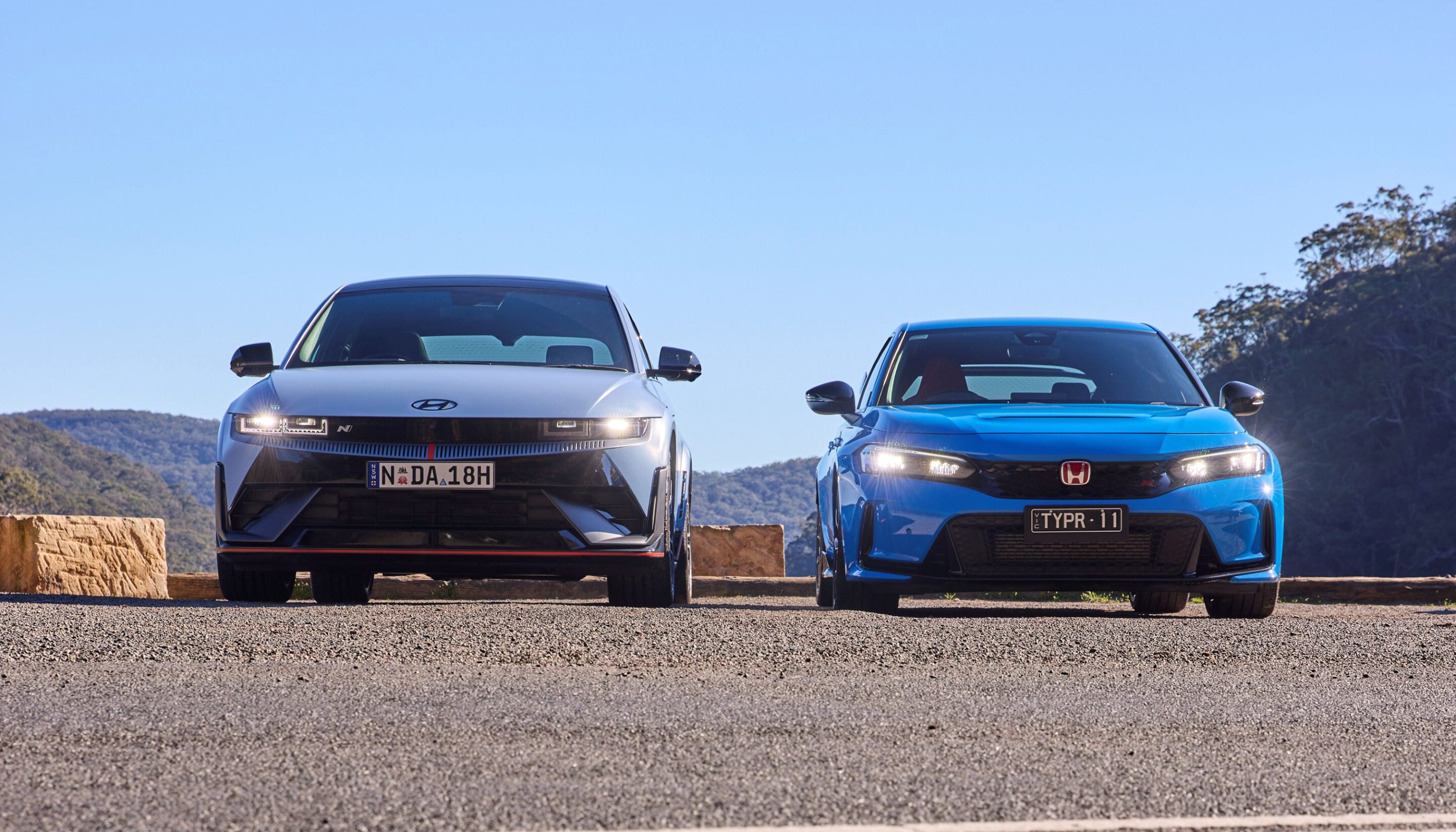 Two modern cars parked side by side on a sunlit road with a clear blue sky behind them. left car is white, right car is blue, with hills in the distance.