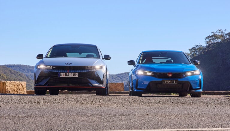 Two modern cars parked side by side on a sunlit road with a clear blue sky behind them. left car is white, right car is blue, with hills in the distance.
