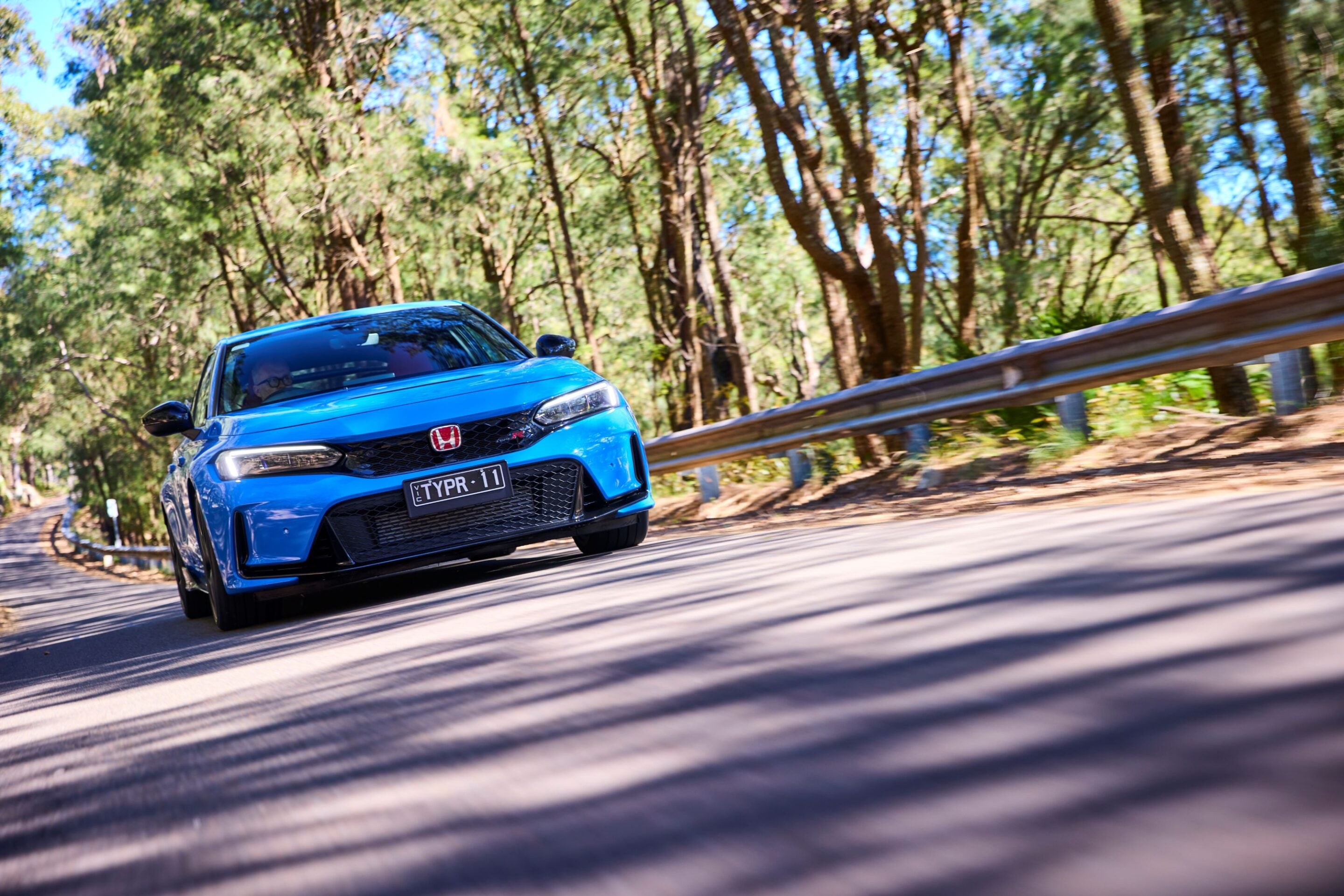 Blue Honda sedan driving quickly along a curved forest road with a metal guardrail and blurred trees in the background.