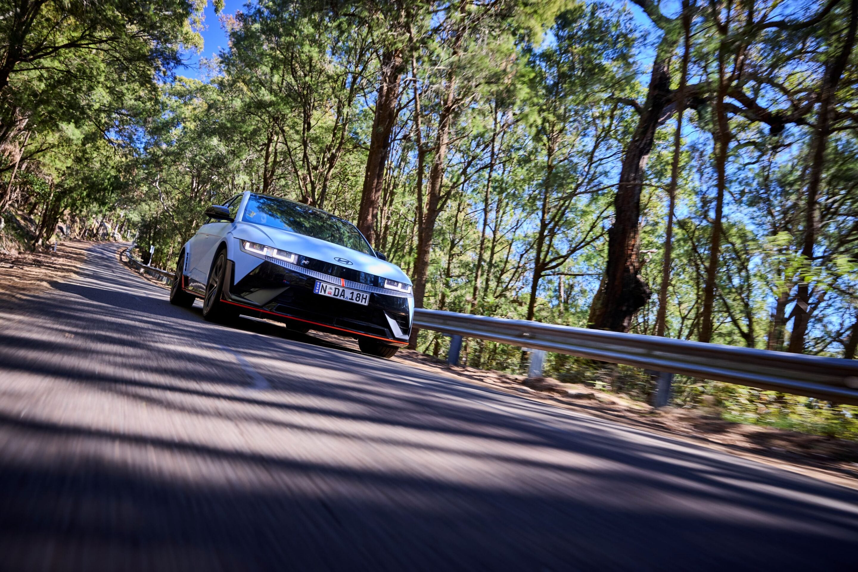White modern sedan driving on a winding forest road, leaves and trees along both sides with a guardrail in the foreground; motion blur suggests speed.