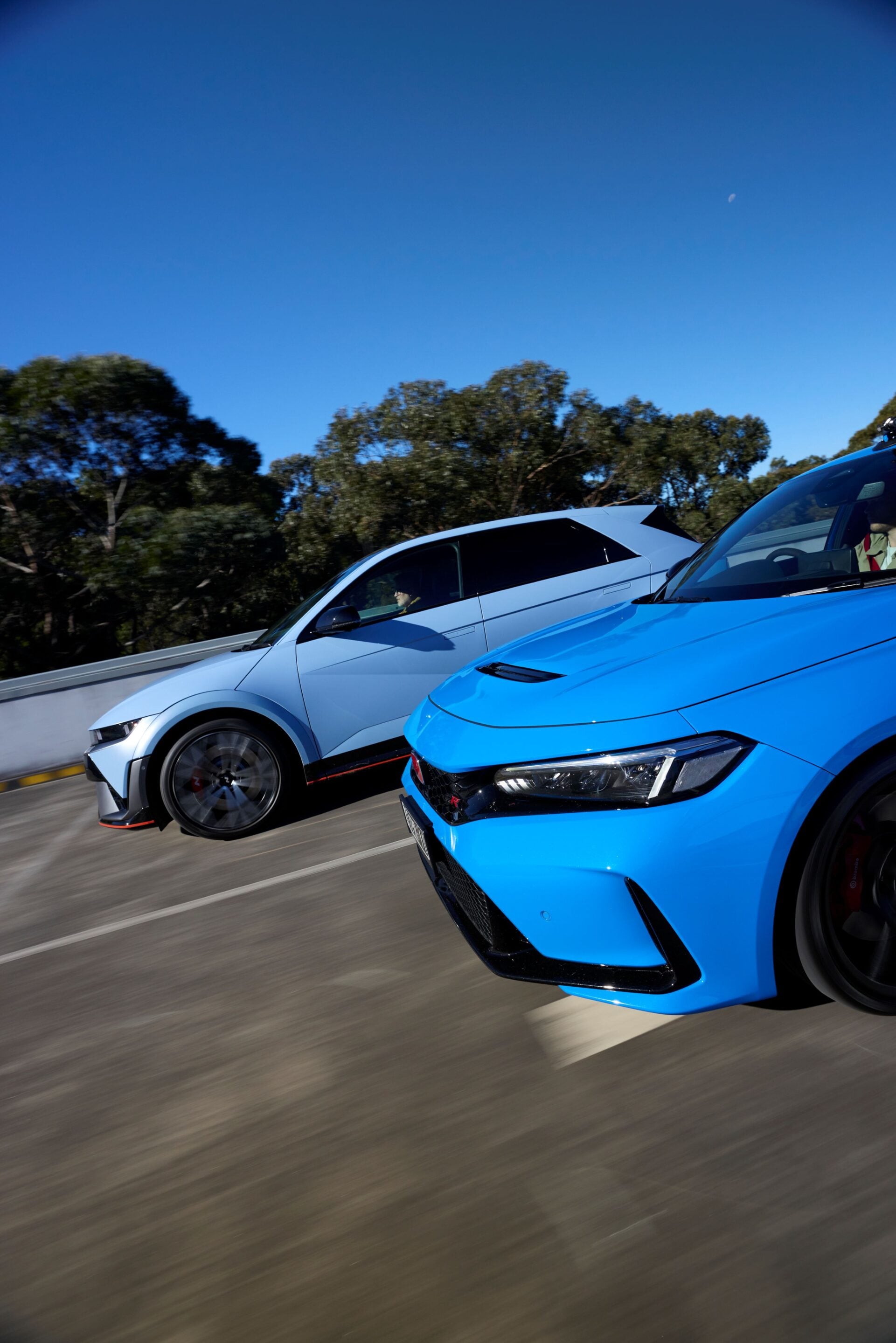 Two modern sports cars, blue and silver, racing on a highway with a clear blue sky and trees in the background.