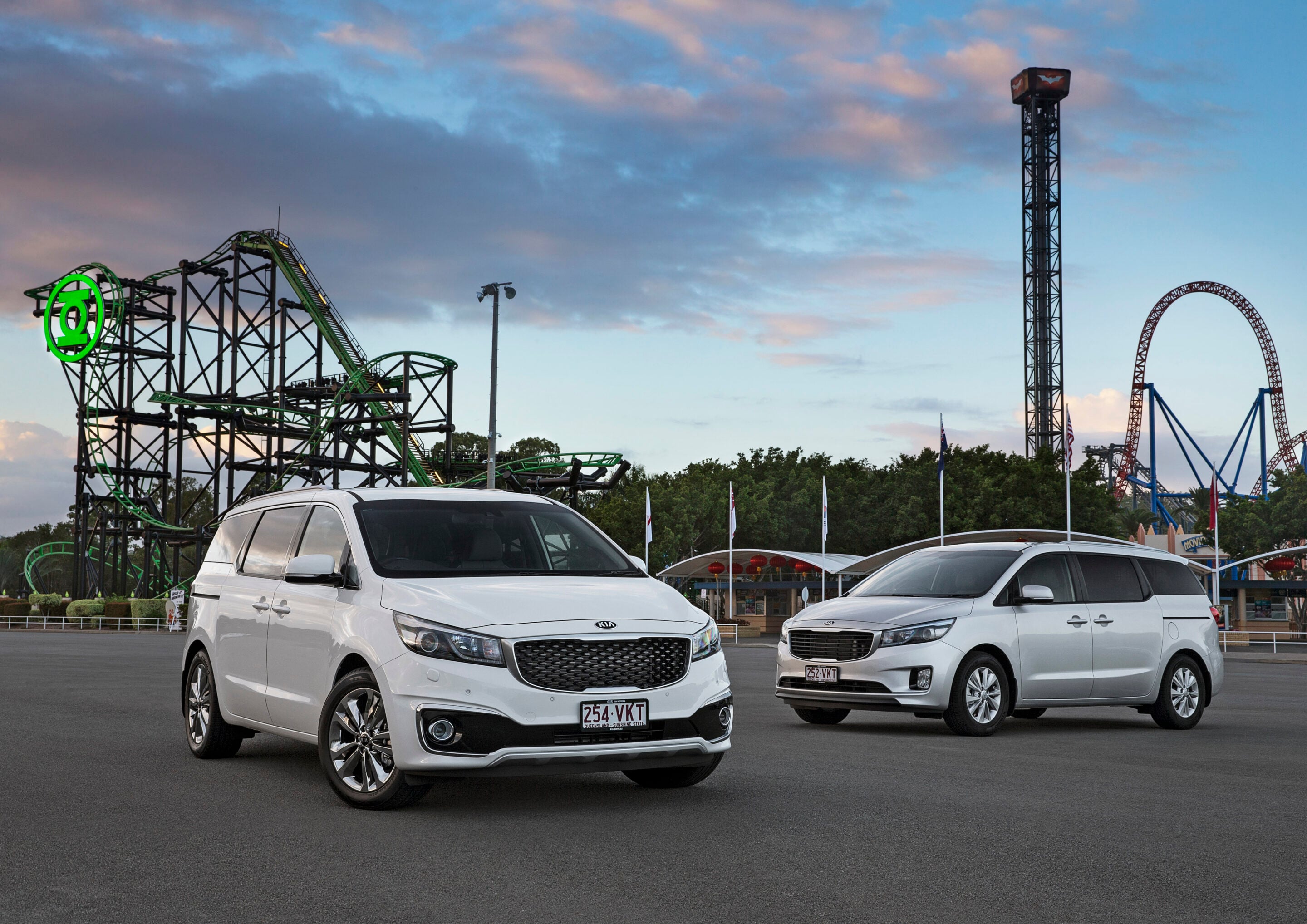 Parking lot in front of an amusement park with white vans and a green roller coaster behind them at sunset/dusk.