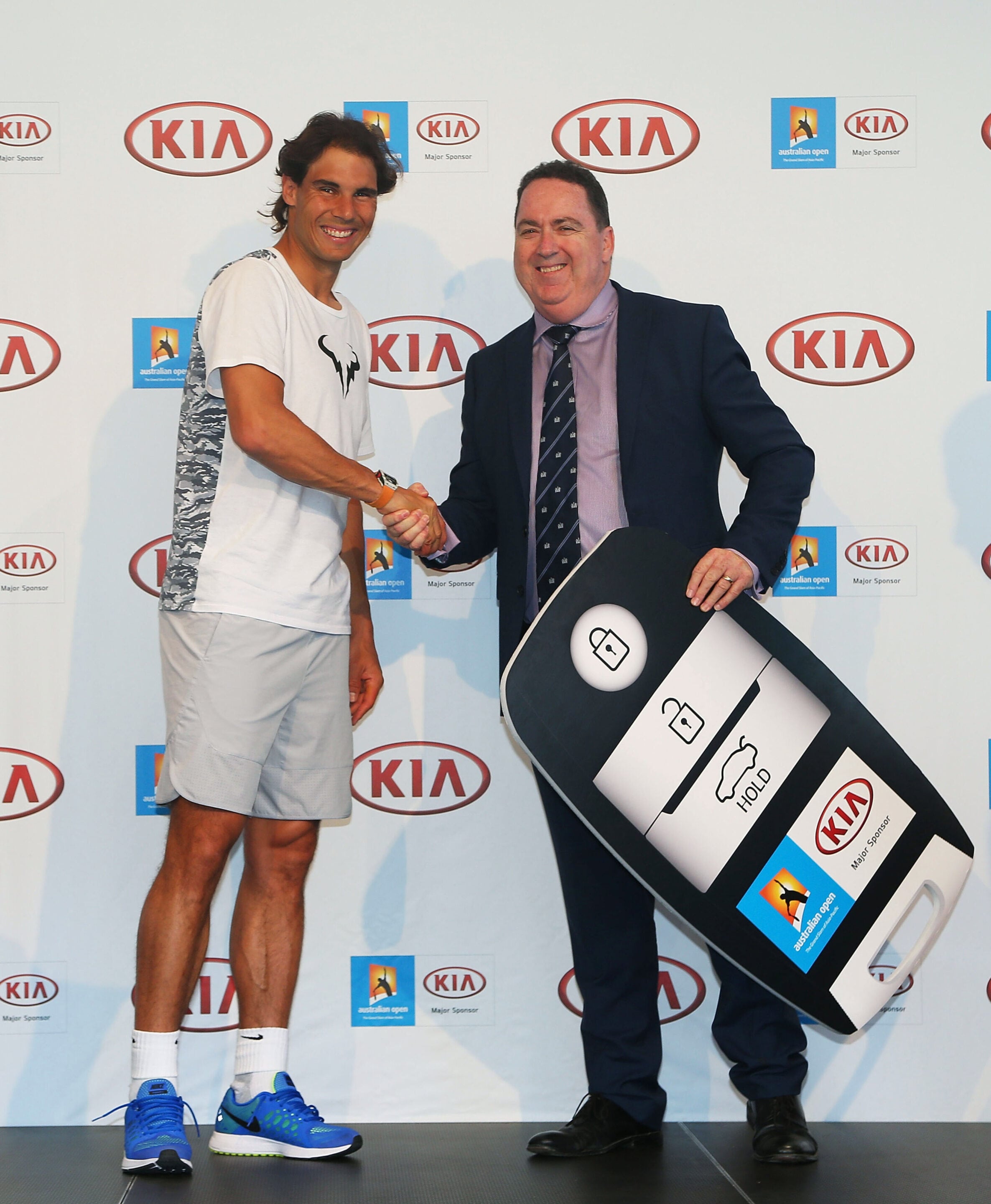 Two men shake hands on a stage with sponsor logos in the background; one in a white T-shirt and blue sneakers, the other in a suit holding a large prize board.