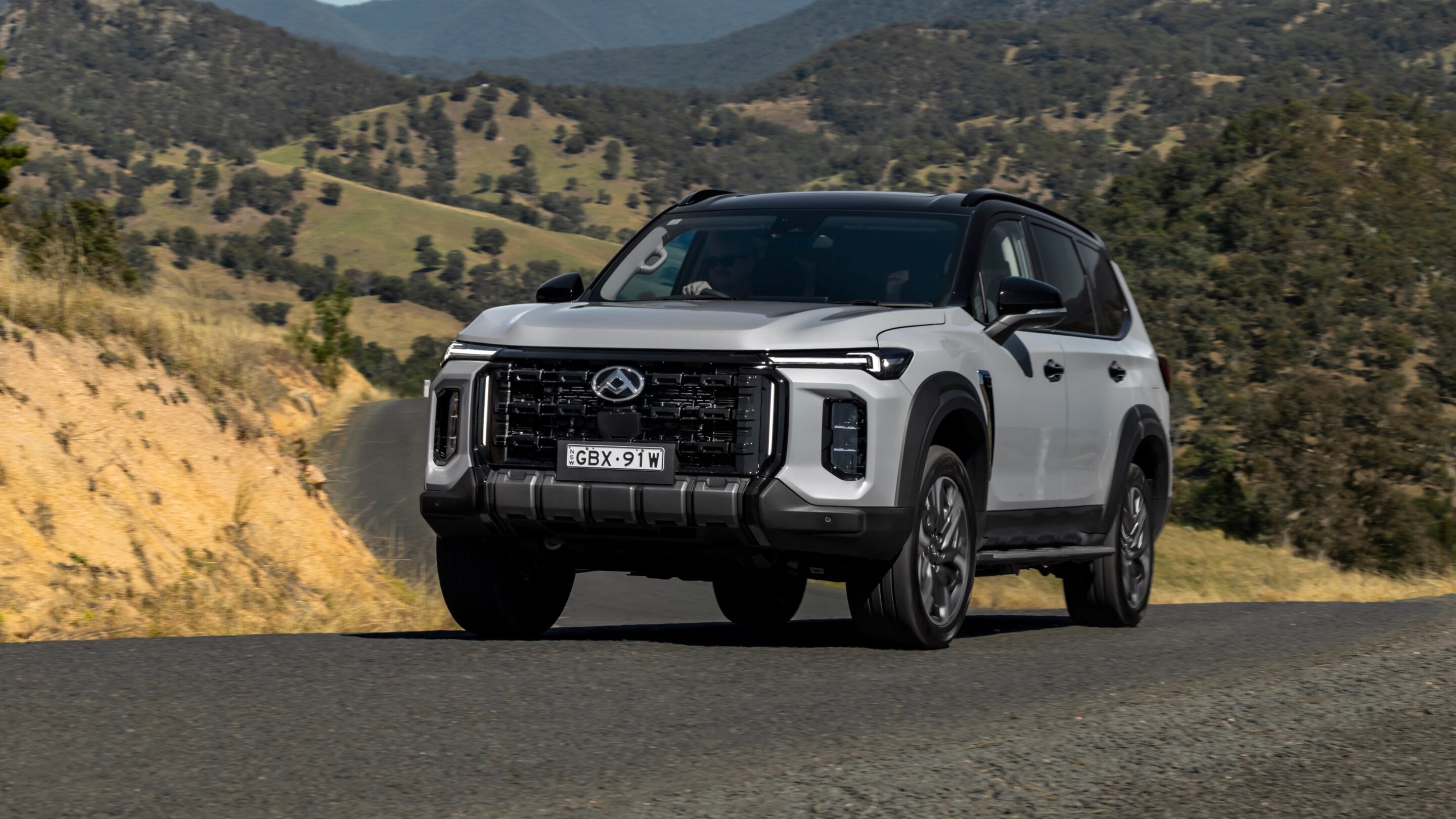 White SUV driving on a winding mountain road with rolling hills in the background.