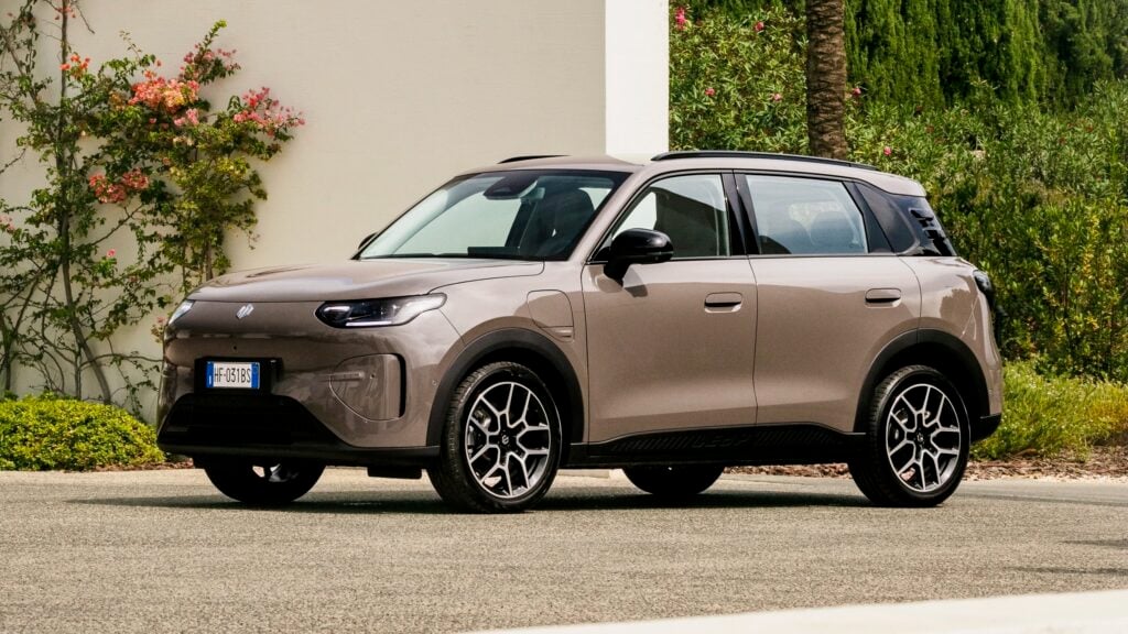Brown compact SUV parked on a street beside a beige wall and green shrubs, shown from the front-left corner with roof rails visible.