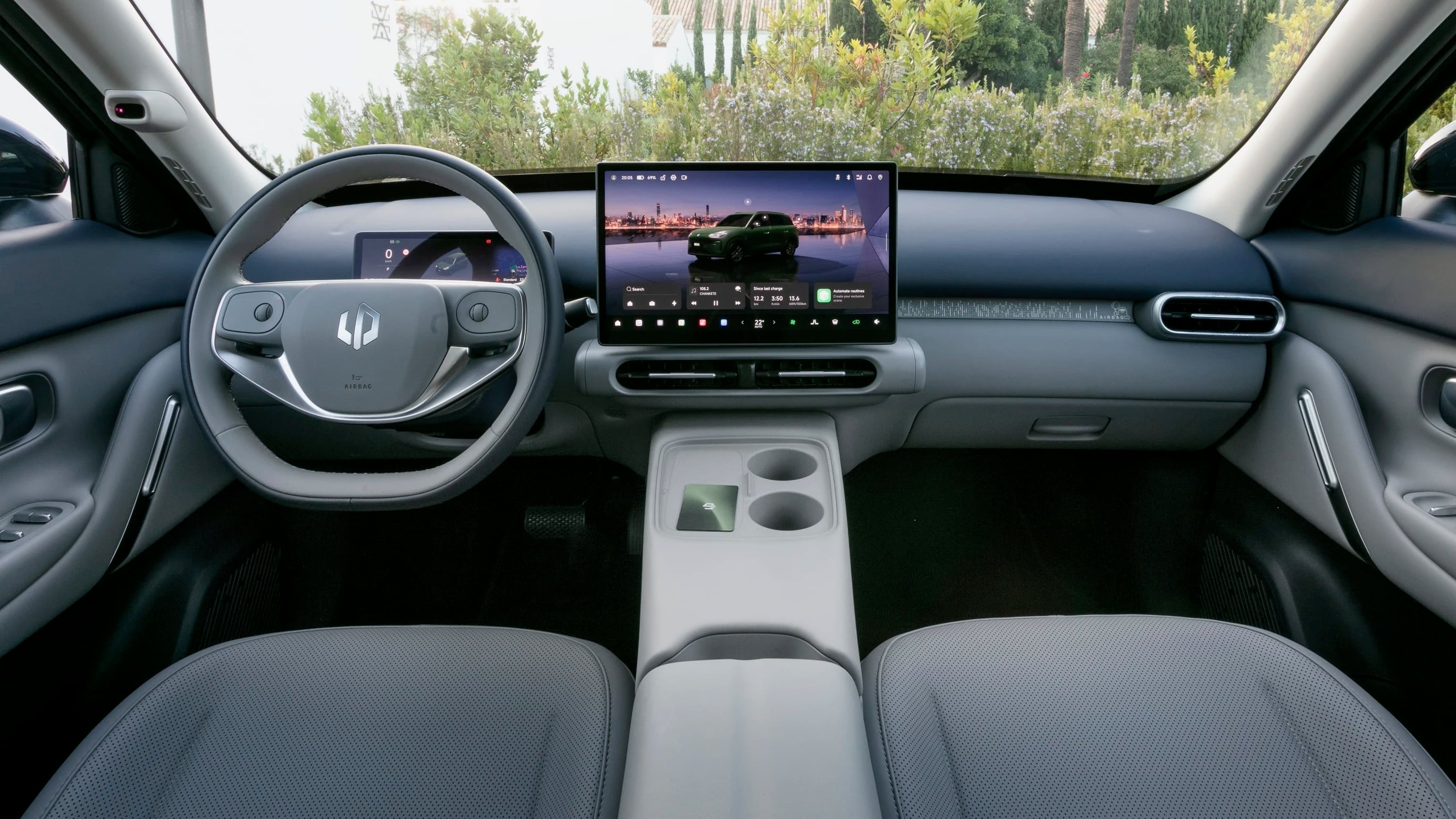 Interior view of a modern car cockpit with a large central touchscreen, steering wheel, and center console; greenery outside through the windshield.