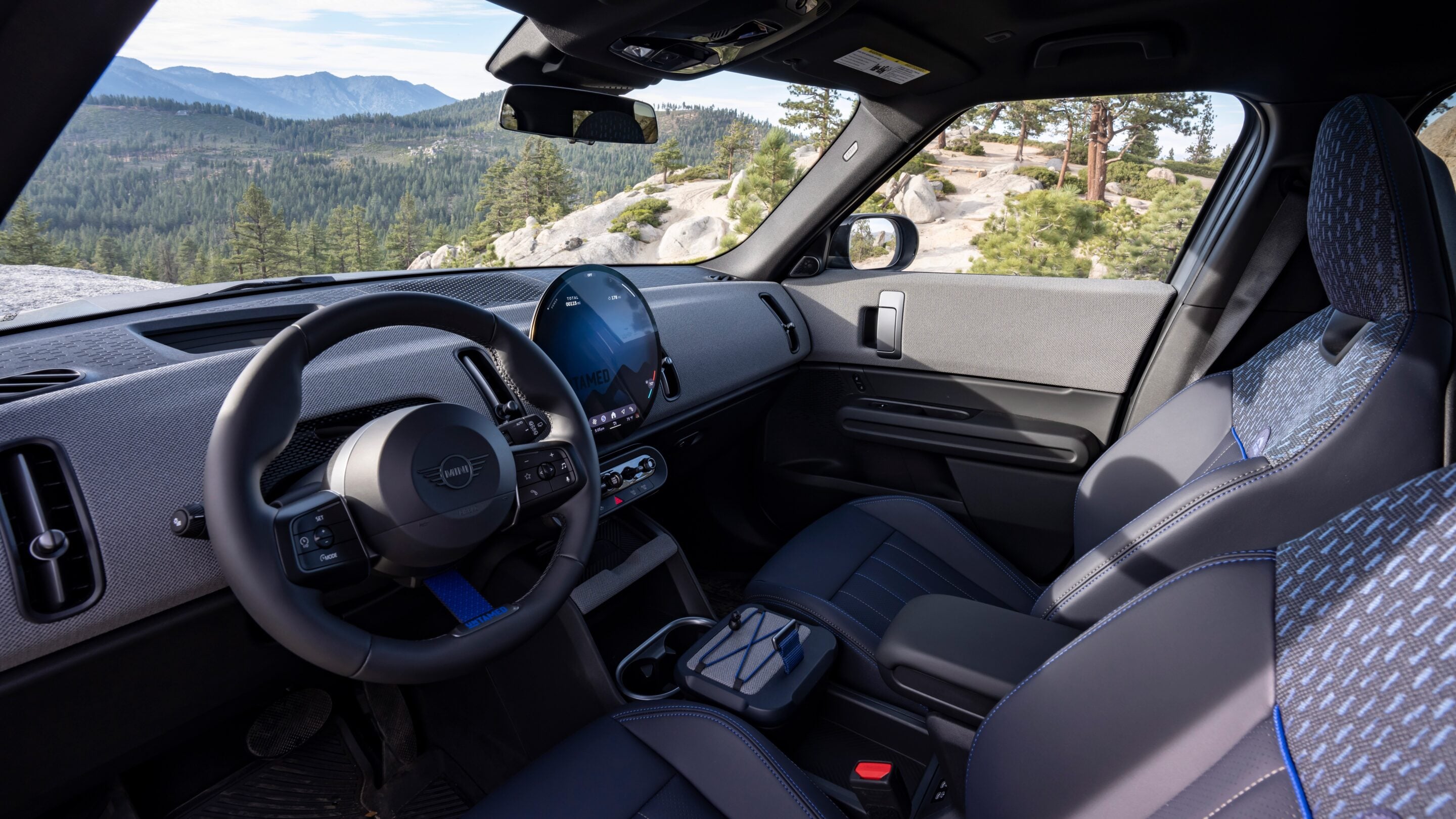 Modern car interior with a digital gauge cluster, steering wheel, and patterned seats; view of rocky, pine-covered landscape outside through the windshield and passenger window.