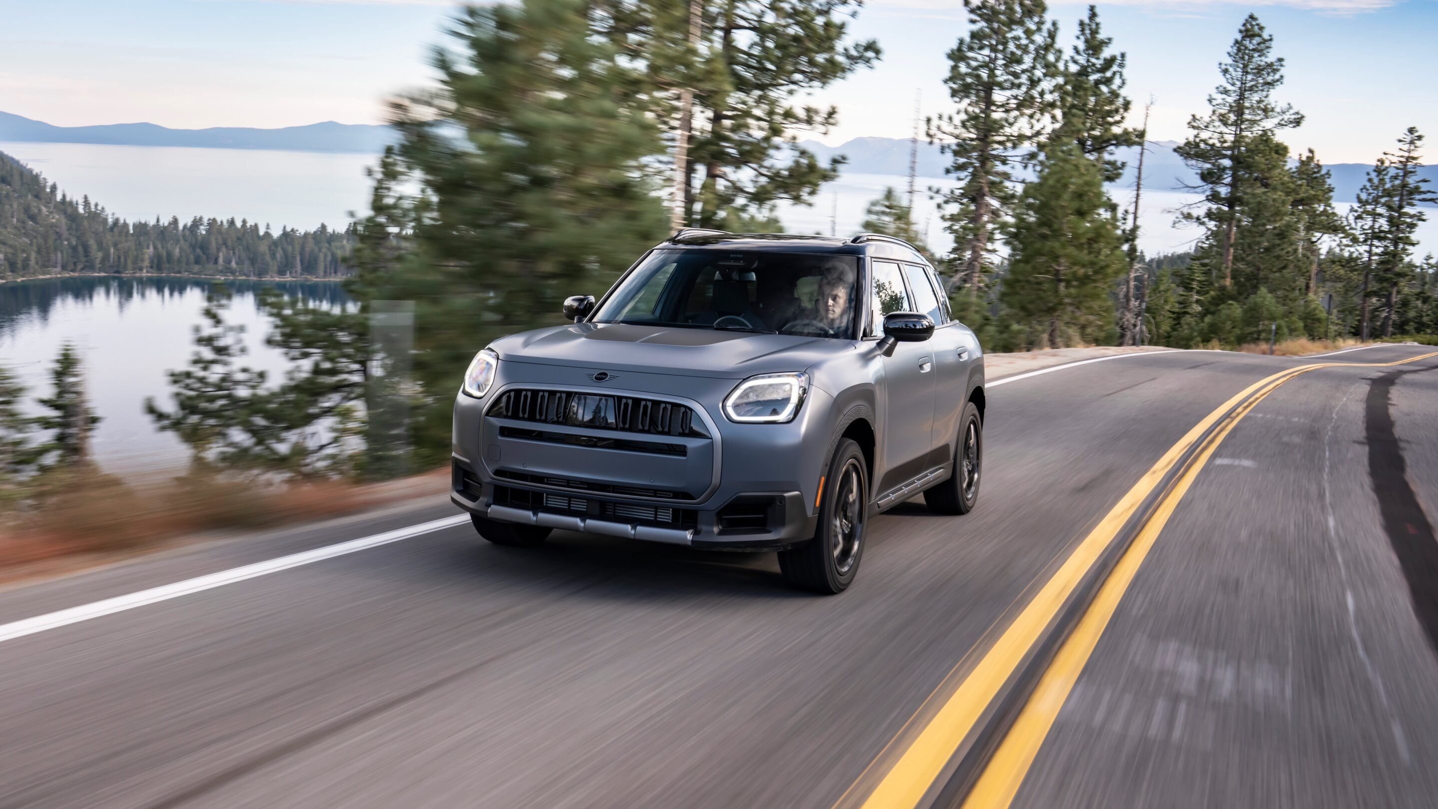 Silver SUV driving along a winding mountain road with a lake and pine forest in the background, motion blurred to show speed.