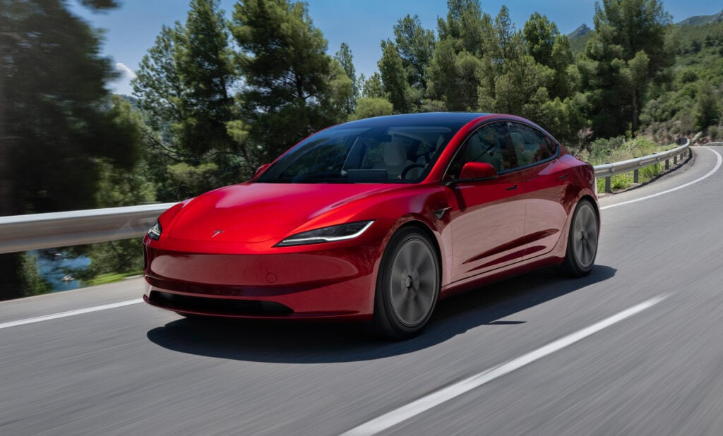 Red Tesla Model 3 driving along a winding mountain road with trees and guardrail in the background.