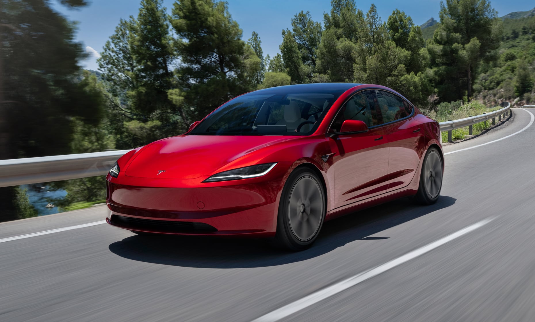 Red Tesla Model 3 driving along a winding mountain road with trees and guardrail in the background.
