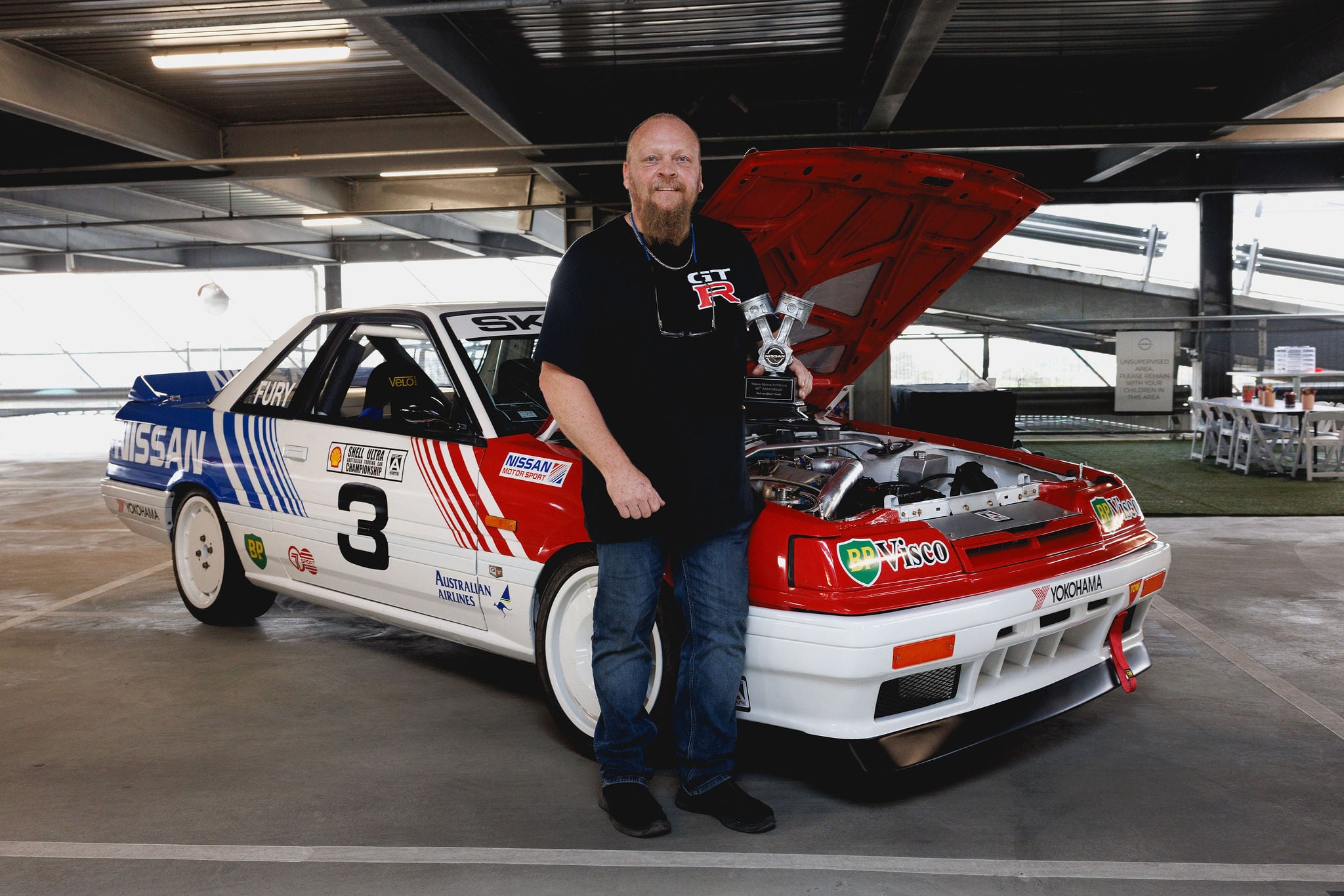 The owner of an R31 race car stands with a trophy alongside his vehicle