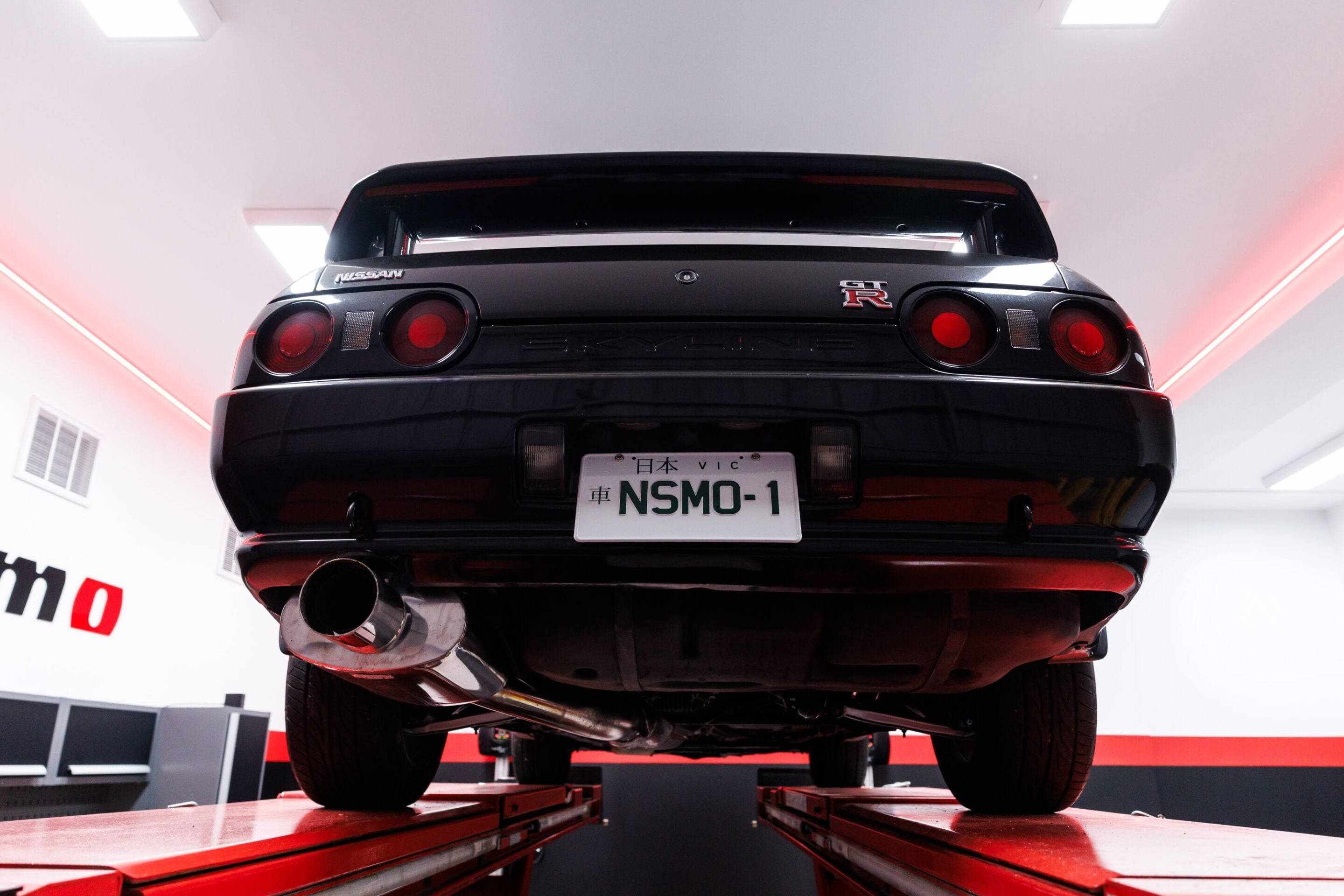 Rear view of a black Nissan GT-R on a car lift, showing red taillights and a European-style license plate NSMO-1 in a bright garage.