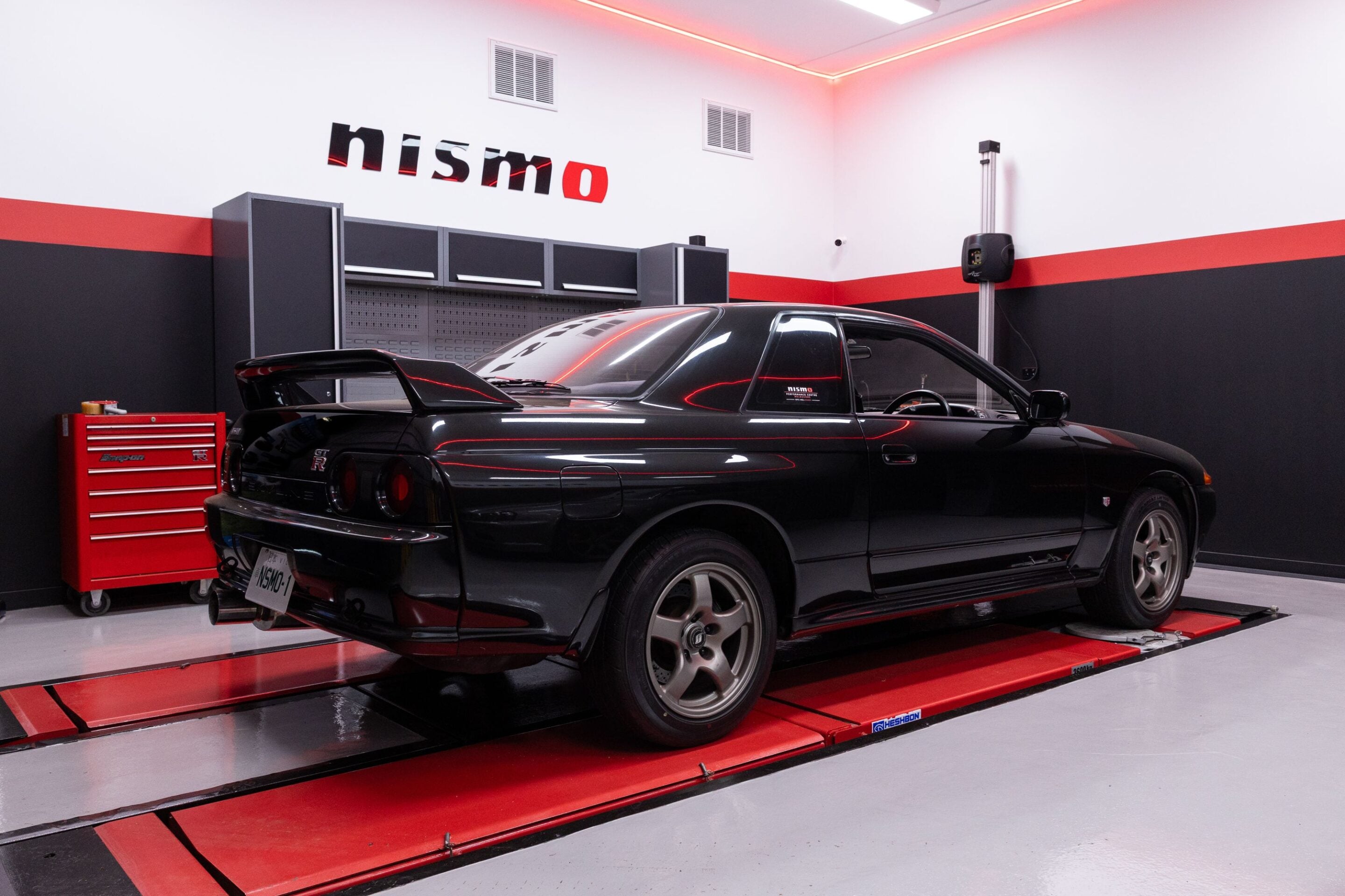 Black Nissan GT-R on a red hydraulic lift in a sleek automotive workshop with gray cabinets and red-accent walls.