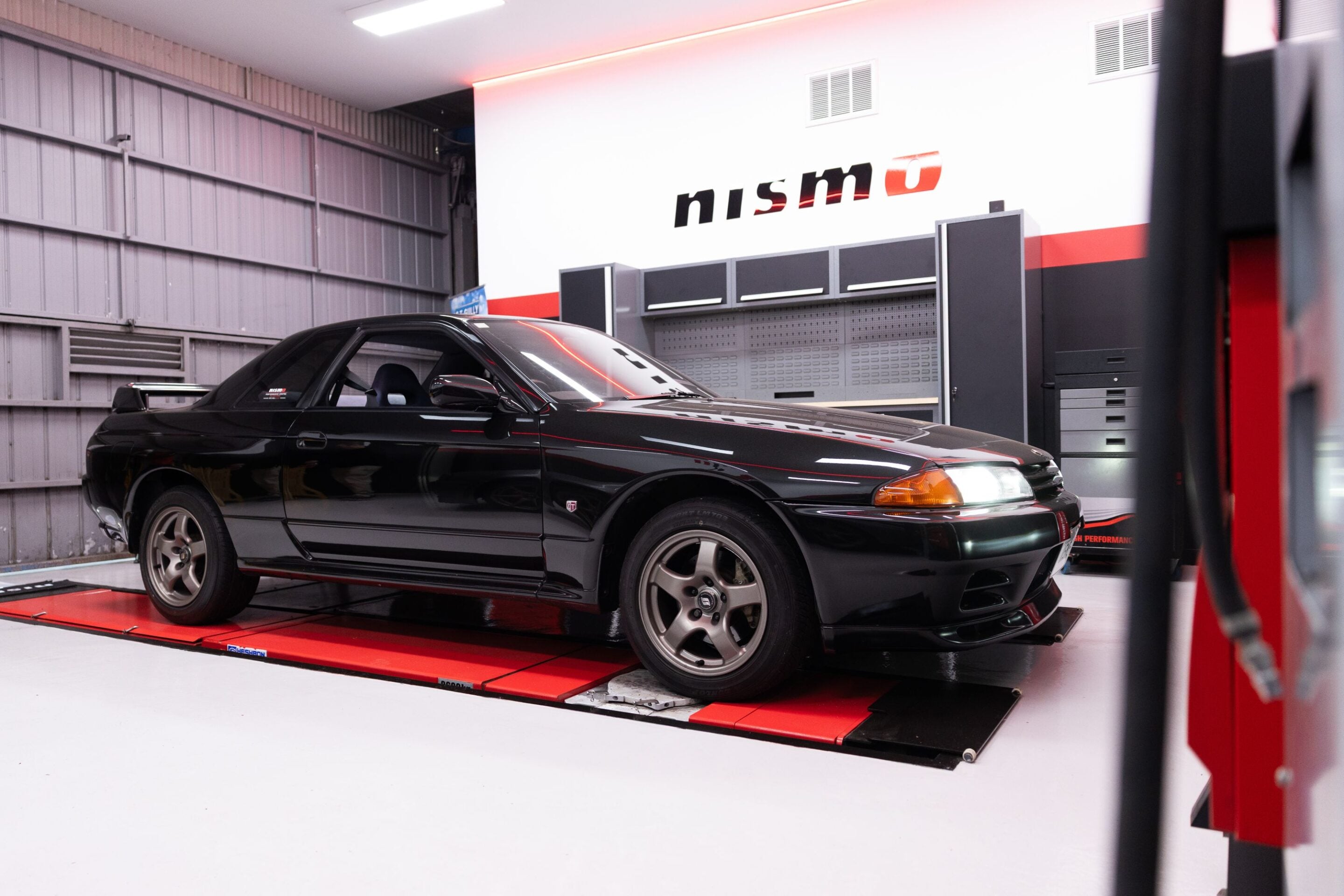 Black sports coupe on a red car lift inside a gray workshop with the Nismo logo on the wall above the cabinets.
