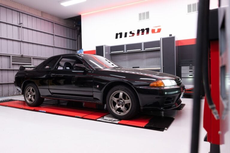 Black sports coupe on a red car lift inside a gray workshop with the Nismo logo on the wall above the cabinets.