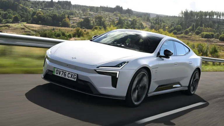 White electric sedan driving on a curved highway with a blurred green countryside backdrop and guardrail on the left. License plate OV76 CXG visible in the front.