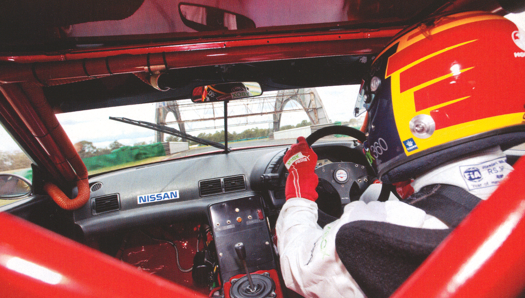Inside a right-hand-drive race car cockpit: driver in a colorful helmet and red racing gloves grips the steering wheel, with a Nissan badge on the dashboard and track visible through the windshield.