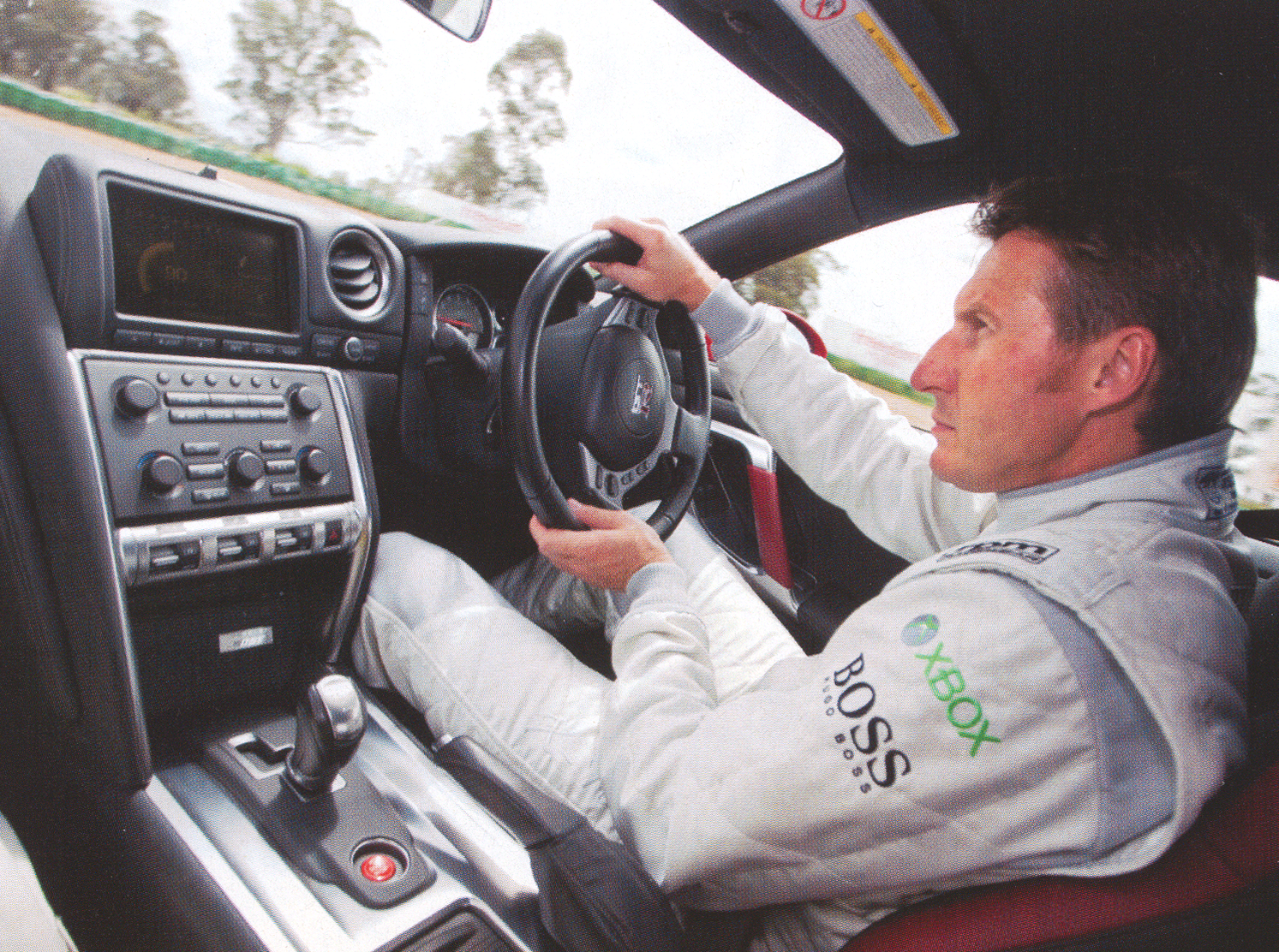 Race car driver in a gray suit grips the steering wheel inside a car cockpit, with dashboard controls nearby