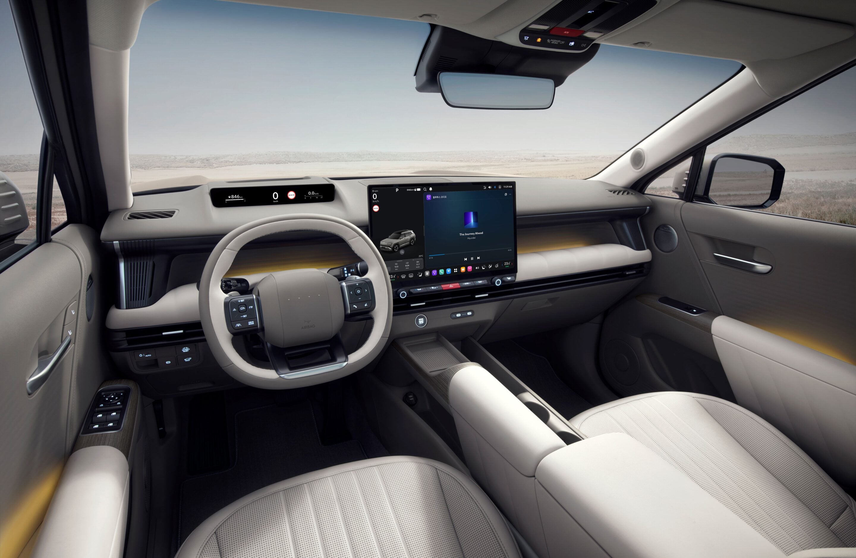 Modern car interior with a large central touchscreen display, digital instrument panel, and light beige upholstery; desert landscape seen through the windshield.
