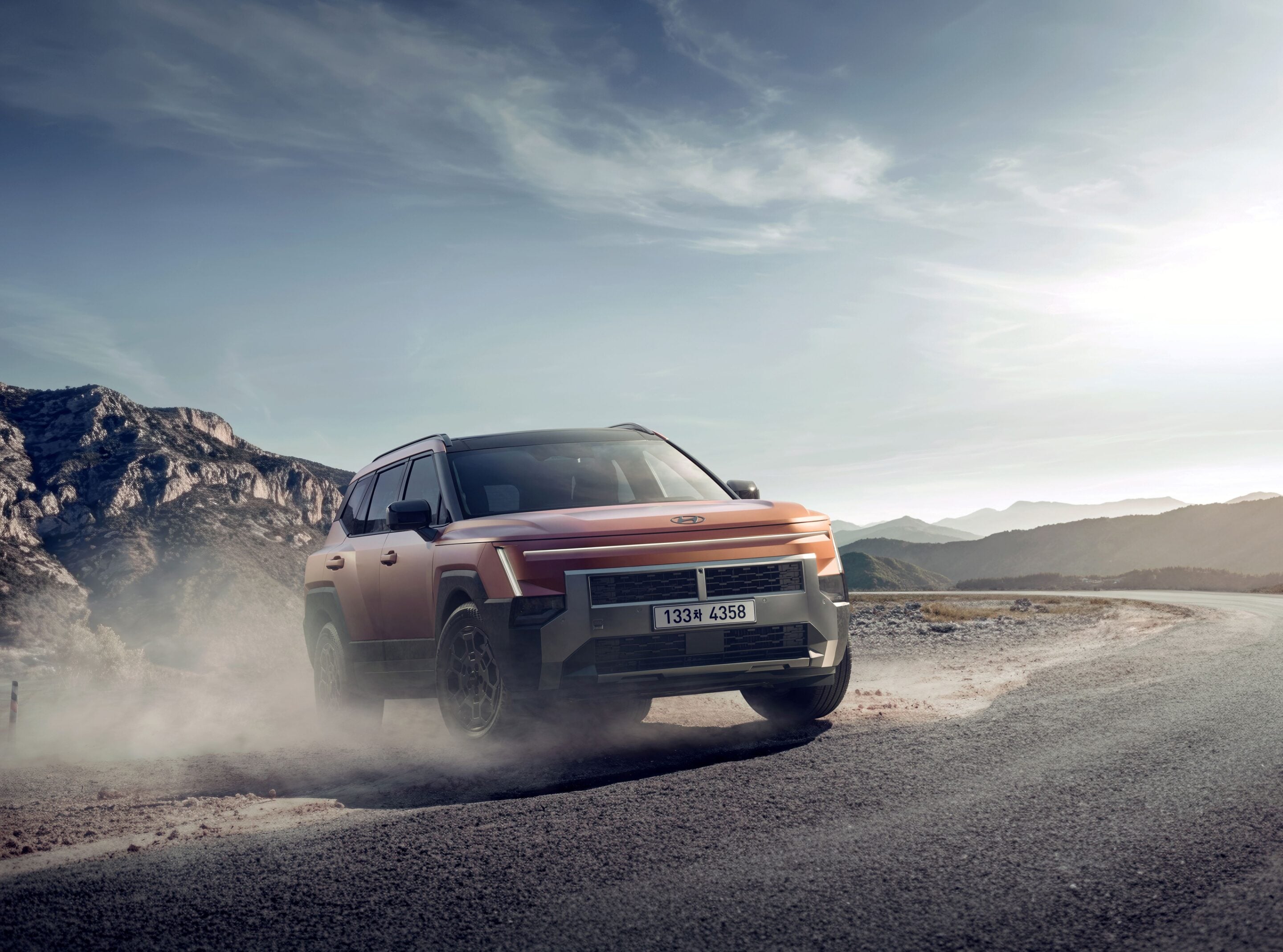 Orange SUV driving on a dusty mountain road with rocky hills and a clear blue sky in the background, clouds above.