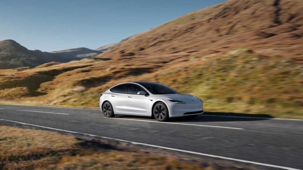 White electric sedan (Tesla) driving along a two-lane mountain road with brown hills in the background, motion blur suggesting speed.