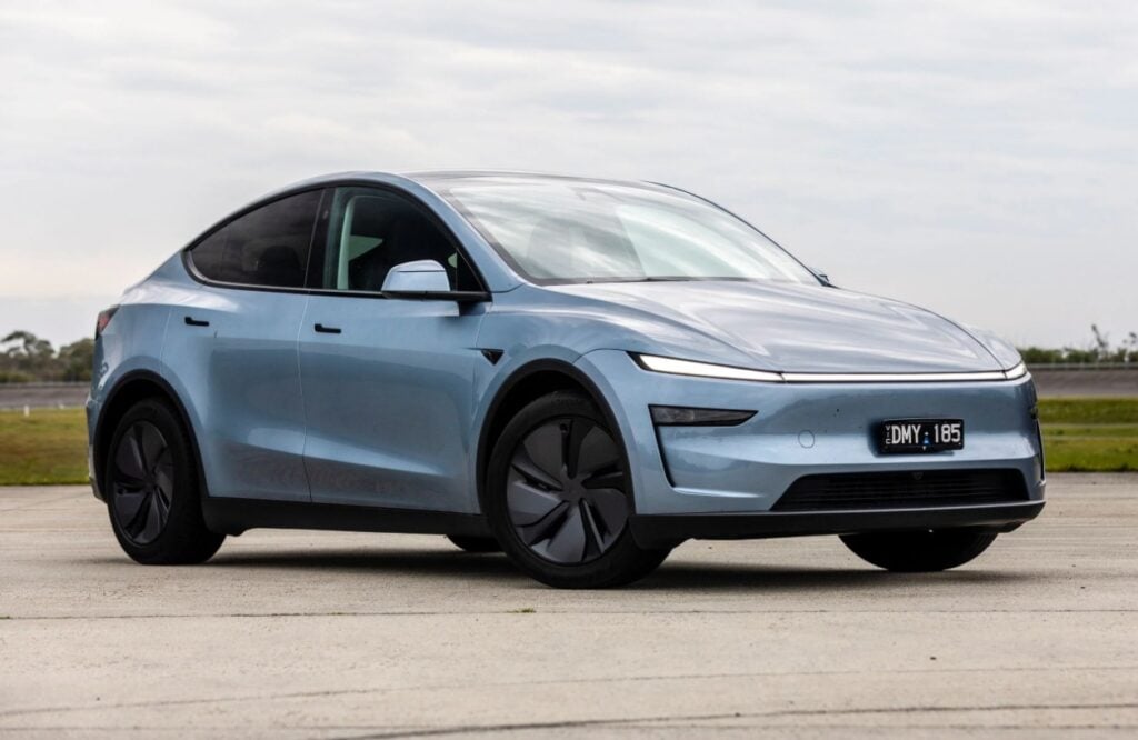 Front-right view of a light blue Tesla Model Y on a wide concrete lot, under a cloudy sky.