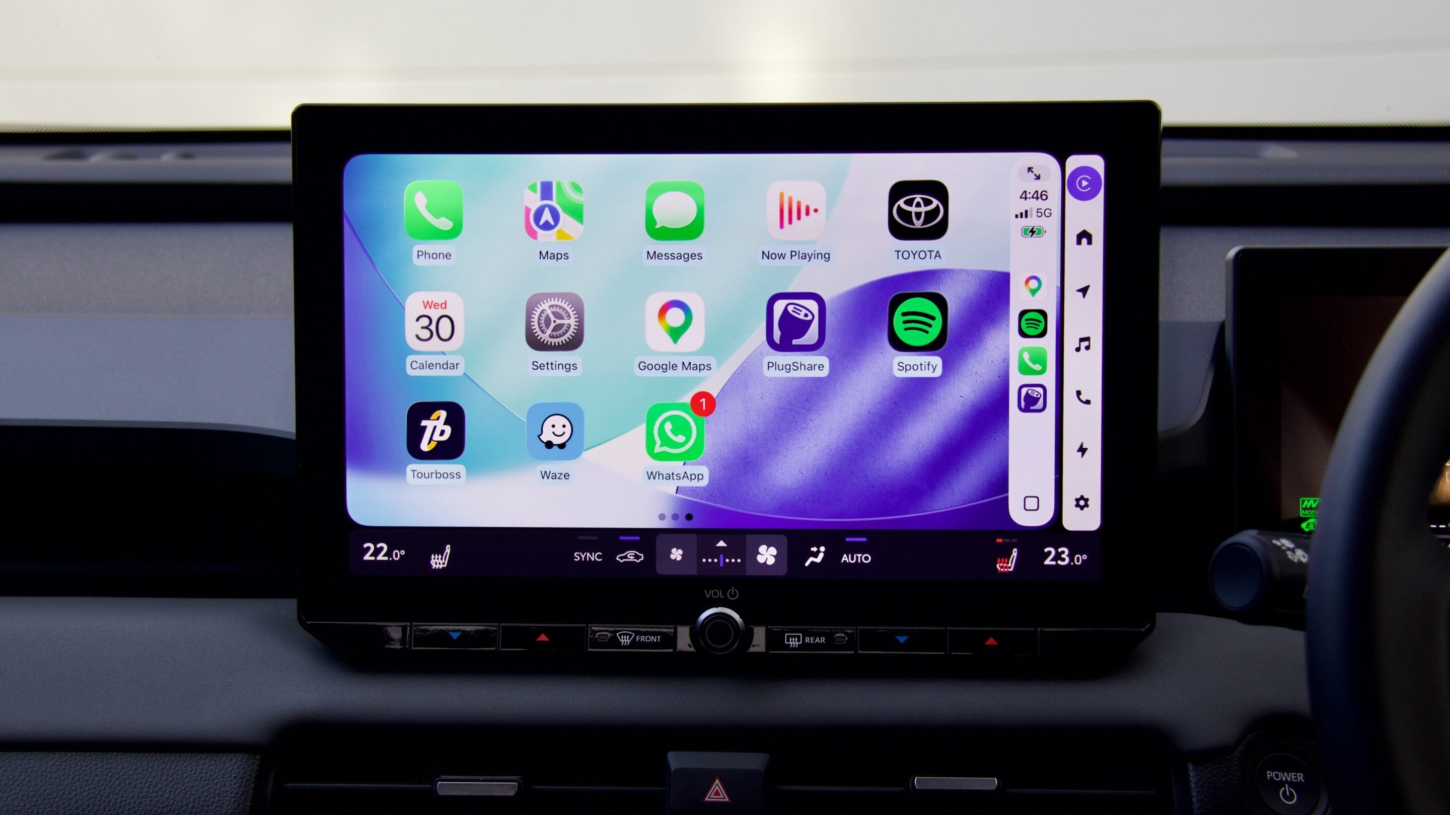 Infotainment touchscreen in a car displaying app icons (Phone, Maps, Messages, Now Playing, Spotify, WhatsApp) on a colorful home screen.