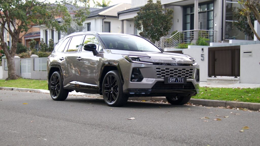 Silver SUV parked on a suburban street in front of modern houses with trees and a gated entrance nearby, during daytime.