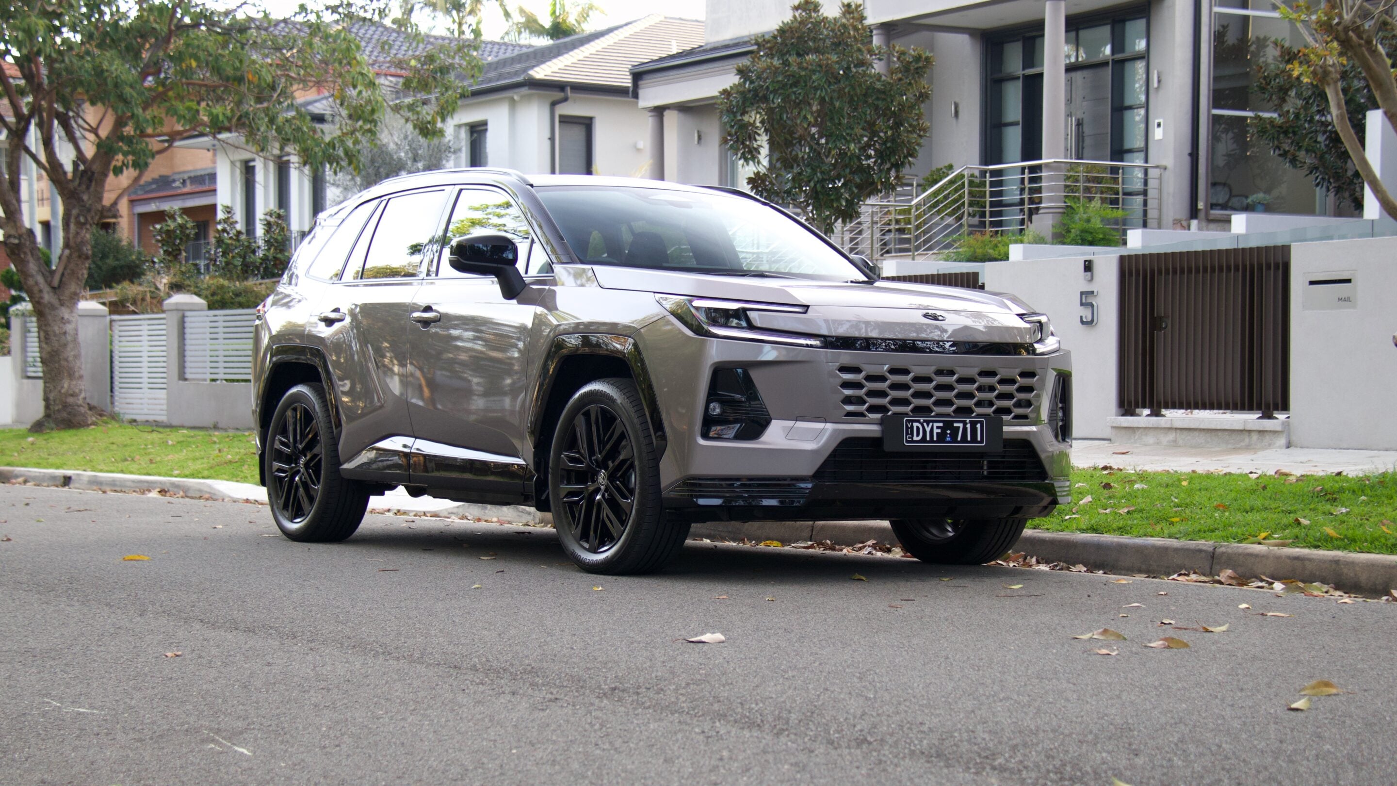 Silver SUV parked on a suburban street in front of modern houses with trees and a gated entrance nearby, during daytime.
