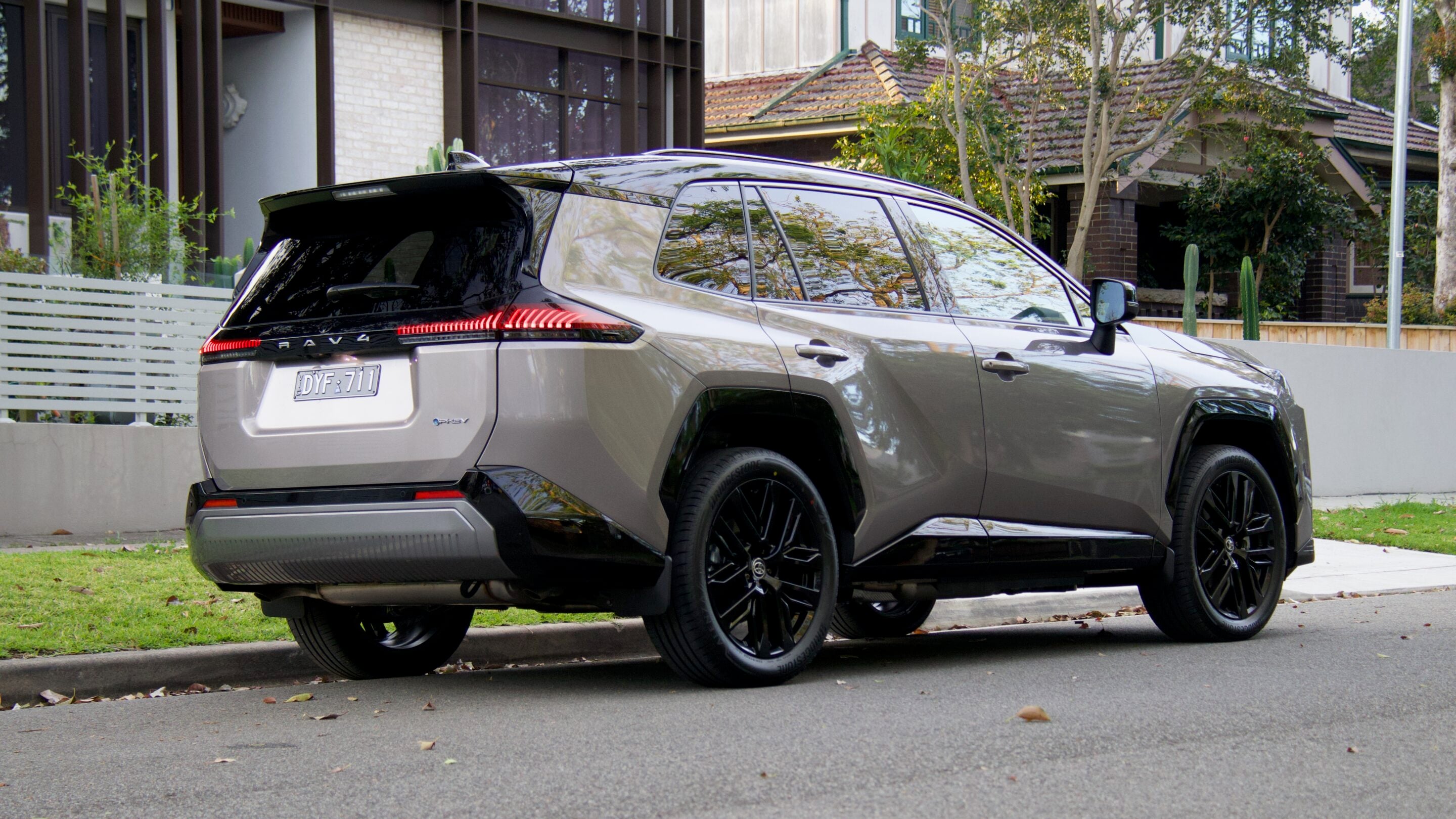 Silver SUV (Toyota RAV4) parked along a suburban street, rear and side view visible with reflections on windows and trees nearby