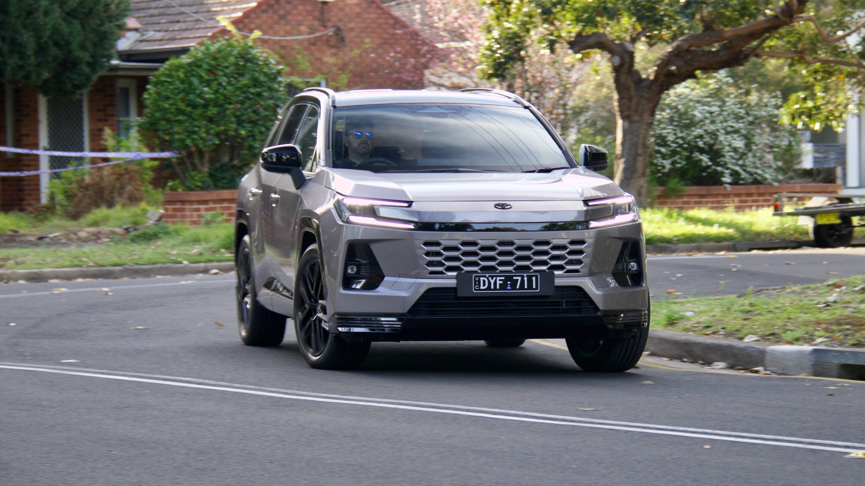 Front view of a silver SUV driving on a suburban street with trees and houses in the background; license plate DYF 711 visible.
