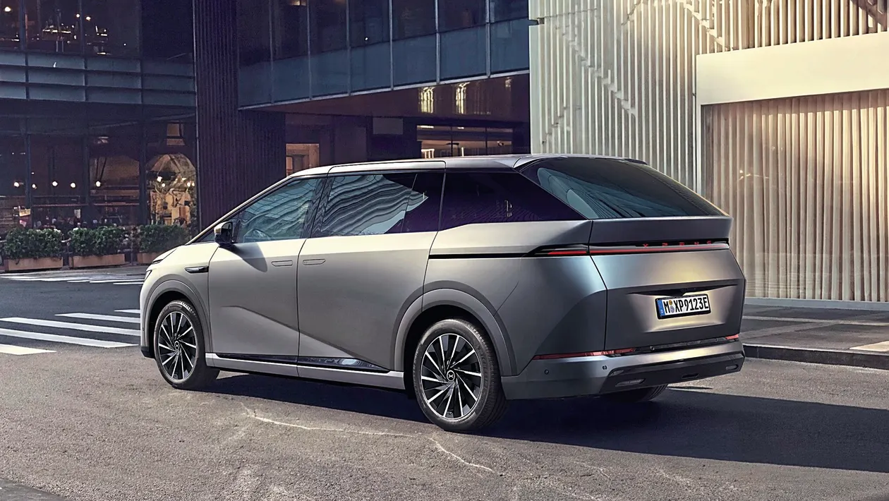 Silver futuristic minivan parked on a city street, featuring a full-width taillight strip and large alloy wheels. The urban backdrop shows glass office buildings and a crosswalk nearby.