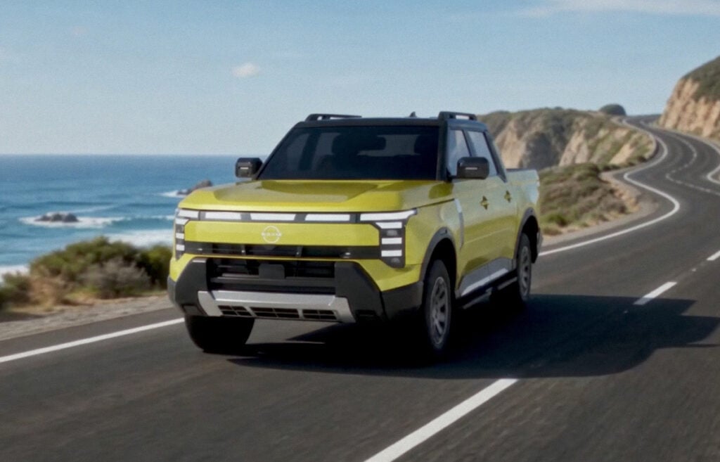 Front three-quarter of a 2027 Nissan Navara Pro PHEV driving on a coastal highway beside ocean cliffs.