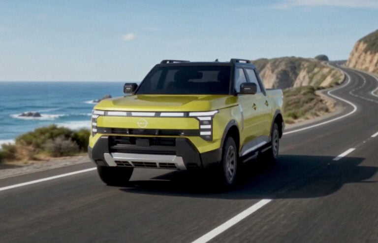 Front three-quarter of a 2027 Nissan Navara Pro PHEV driving on a coastal highway beside ocean cliffs.