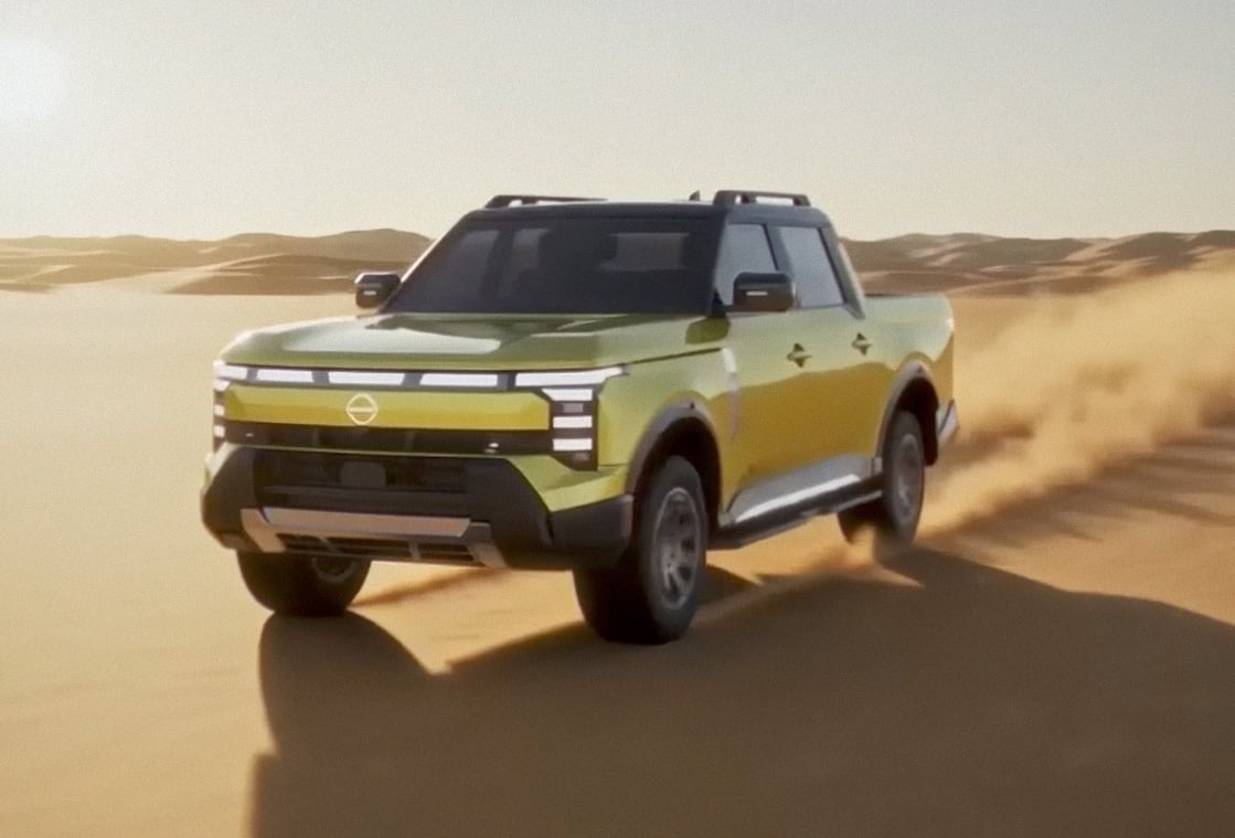 Front three-quarter of a 2027 Nissan Navara Pro PHEV driving across sand dunes with a trail of dust behind the vehicle.
