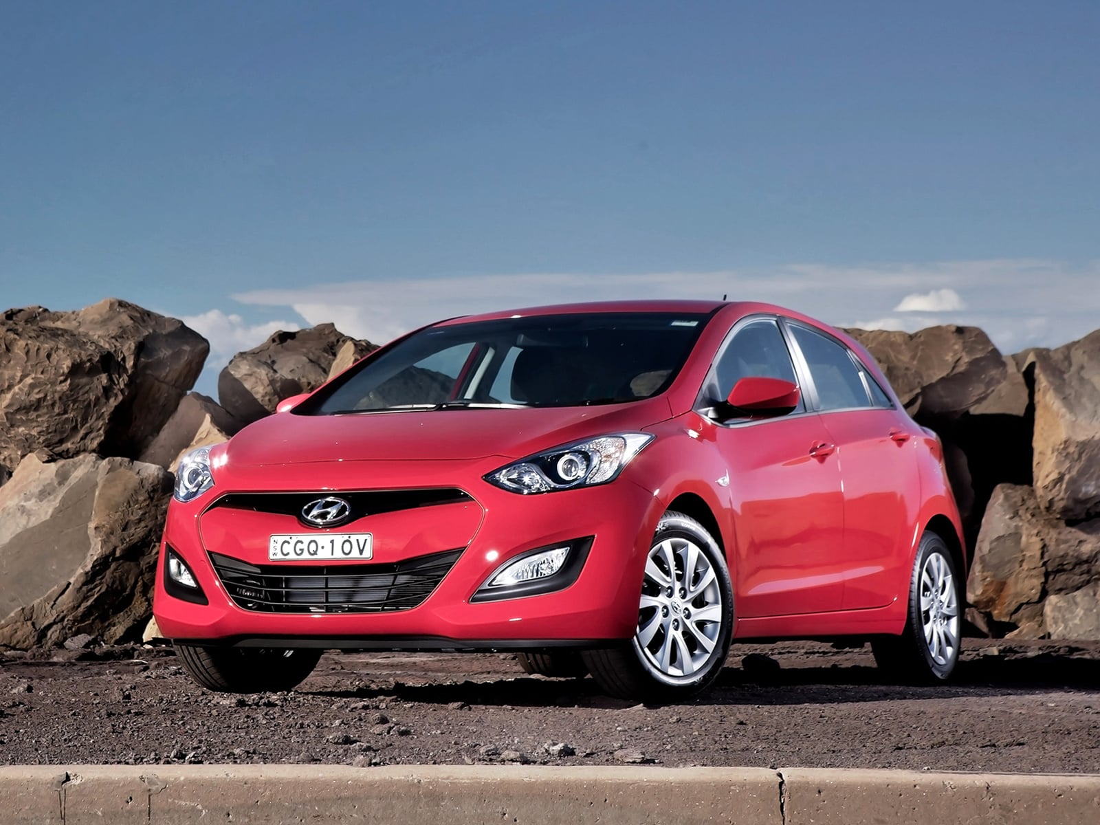 Front three-quarter view of a red Hyundai sedan parked beside large rocks under a clear blue sky.